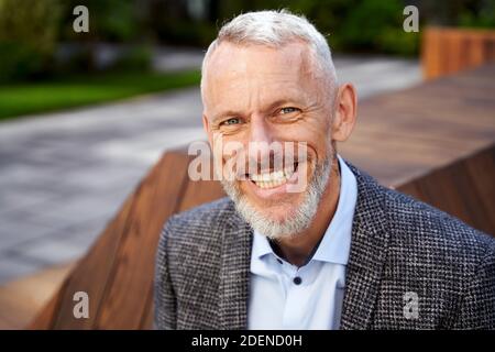 Primo piano ritratto di un elegante uomo d'affari maturo che sorride alla macchina fotografica, seduto sulla panchina all'aperto Foto Stock