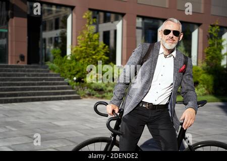 Commuter. Uomo d'affari di media età sicuro ed elegante in occhiali da sole in piedi con la sua bicicletta vicino al suo posto di lavoro e guardando la fotocamera Foto Stock