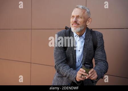 Uomo d'affari moderno di mezza età che guarda premuroso mentre in piedi all'aperto con scooter elettrico e una tazza di caffè Foto Stock