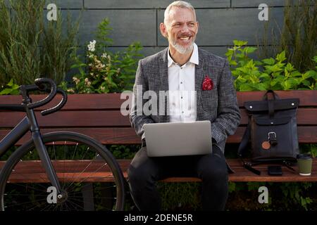 Lavorare all'esterno. Gioioso uomo d'affari moderno di mezza età in abito elegante che lavora sul suo computer portatile mentre si siede sulla panchina all'aperto con una bicicletta accanto Foto Stock