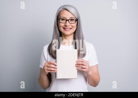 Ritratto fotografico della vecchia donna che mostra un notebook con foglio vuoto occhiali sorridenti isolati su sfondo grigio con copyspace Foto Stock