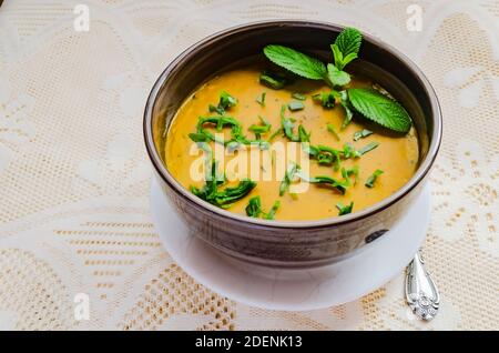 Su uno sfondo lacey si trova una ciotola a strisce marroni di zuppa di zucca densa e senza latte, guarnita con spinaci verdi e menta. Foto Stock