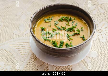 Su uno sfondo lacey si trova una ciotola a strisce marroni di zuppa di zucca densa e senza latte, guarnita con spinaci verdi. Foto Stock