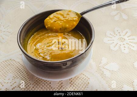 Un cucchiaio sta sollevando la zuppa di zucca liscia e spessa da una ciotola che si trova in un piccolo piatto bianco su una tovaglia di pizzo. Foto Stock