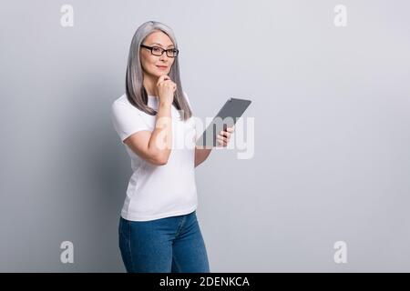 Foto di intelligente donna senior indossare occhiali casual outfit reggendo moderno braccio di gadget mento isolato sfondo grigio Foto Stock