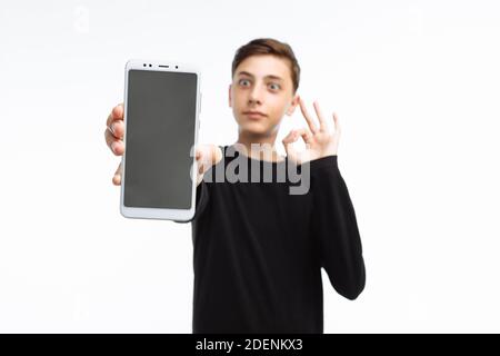 Ritratto di un adolescente, un ragazzo che tiene un telefono con schermo bianco, e mostra un dito sullo schermo, in una t-shirt nera, su uno sfondo bianco, Foto Stock