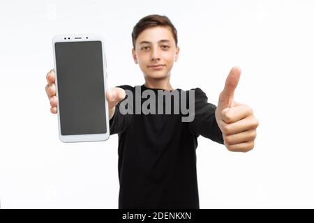 Ritratto di un adolescente, un ragazzo che tiene un telefono con schermo bianco, e mostra una classe di dita, in una t-shirt nera, su uno sfondo bianco, Foto Stock