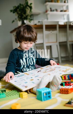 Il ragazzino tiene un libro e vede i disegni. Il bambino si siede sul pavimento. Vicino giocattoli sparsi. Il concetto di famiglia Foto Stock