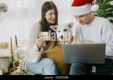 Felici i giovani con gli animali domestici che acquistano online o che chattano in videochiamata con il computer portatile nella sala festiva del boho. Coppia elegante con carino cane bianco in luci e. Foto Stock