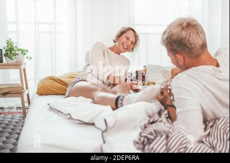 Ridendo capelli biondi giovane coppia di adulti in pigiama pigro rilassante sdraiato in un letto accogliente nella camera da letto e di avere una chiacchierata tranquillamente spensierata. Coppia Foto Stock