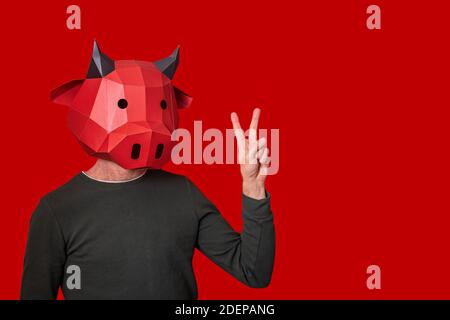 Giovane uomo in rosso maschera 3d di mucca toro sulla testa su uno sfondo isolato. Sta mostrando il segno di pace con gesti. Vuoto per banner, template, pl Foto Stock