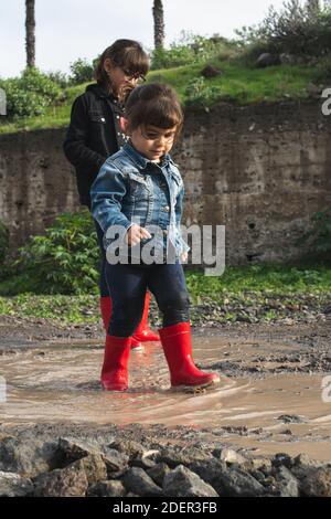 Ragazza che salta in pudddddle fangose indossando i wellies rossi, riempiendo in su di fango Foto Stock