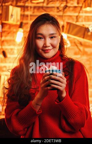 Ragazza asiatica in maglione con tazza di cacao caldo. Caffè con marshmallows e lollipop. Atmosfera natalizia. Umore di nuovo anno. Ritratto romantico. Lampadine e presenti scatole sfondo di muro di mattoni. Foto Stock