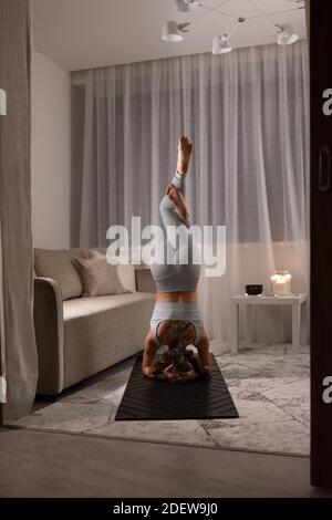 Vista posteriore di una donna anonima che fa supportato Headstand con Eagle Gambe a casa Foto Stock