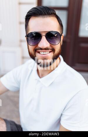 Elegante hipster in una t-shirt bianca con occhiali da sole e una barba, in piedi vicino a una parete di legno bianco Foto Stock