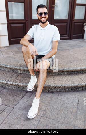 Elegante hipster in una t-shirt bianca con occhiali da sole e una barba, seduto sui gradini vicino all'edificio bianco Foto Stock