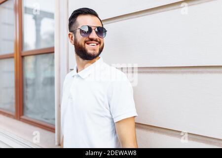 Elegante hipster in una t-shirt bianca con occhiali da sole e una barba, in piedi vicino a una parete di legno bianco Foto Stock