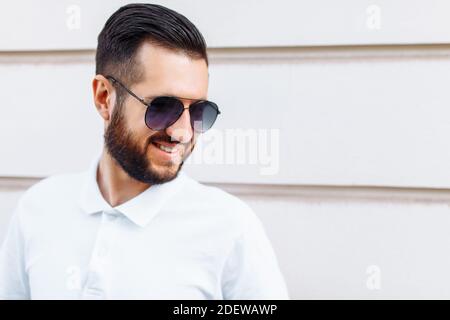 Elegante hipster in una t-shirt bianca con occhiali da sole e una barba, in piedi vicino a una parete di legno bianco Foto Stock