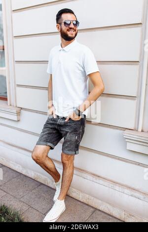 Elegante hipster in una t-shirt bianca con occhiali da sole e una barba, in piedi vicino a una parete di legno bianco Foto Stock