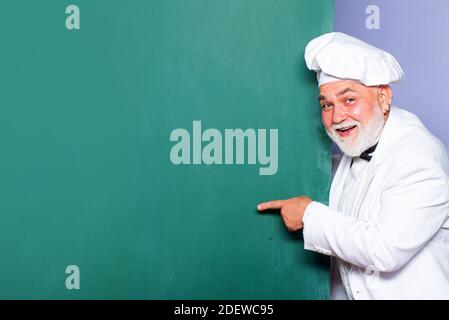 Capo cuoco senior. Ritratto del capo cuoco con il cartello a mano sullo sfondo della lavagna. Cuoco uomo eccitato che indossa uniforme mostrando testo copy space. Foto Stock