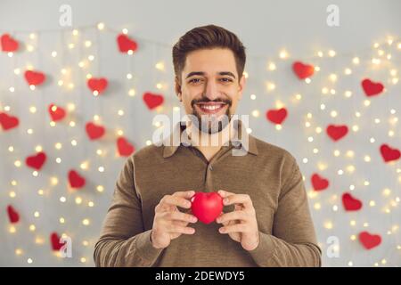 Ritratto di un giovane sorridente che tiene un piccolo cuore rosso di San Valentino e guardando la fotocamera Foto Stock