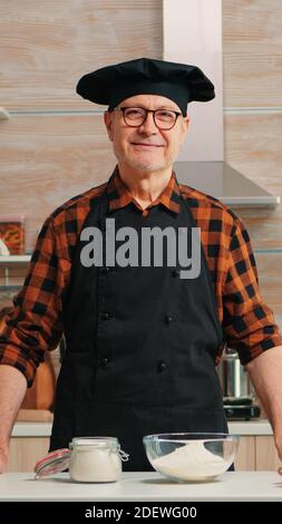Ritratto dello chef che indossa bonete guardando la fotocamera e sorridendo. Panettiere anziani in cucina uniforme preparare ingredienti di pasticceria su tavola di legno pronti a cucinare pane, torte e pasta gustosi fatti in casa Foto Stock