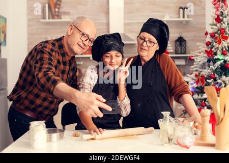 Nonno e bambino che assumono selfie il giorno di natale indossando zafferano e bonette. Felice allegra joyfull adolescente ragazza aiutare donna anziana preparare dolci biscotti per celebrare le vacanze invernali. Foto Stock