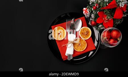 Impostazione per la cena di Natale di festa su tavolo nero con decorazione di Capodanno e arance secche. Palline rosse di Natale in un bicchiere di vino Foto Stock