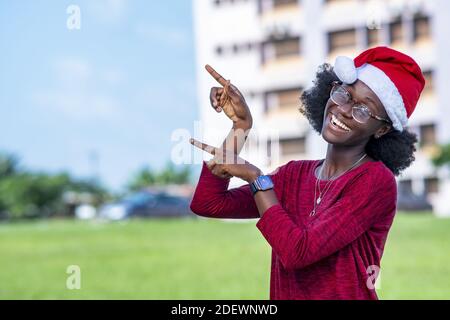 Una bella donna nera sorridente in un cappello di Santa e gli occhiali che puntano le dita verso il cielo Foto Stock