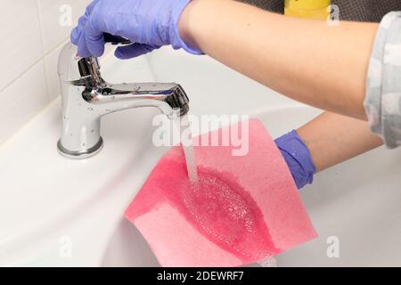 la mano femminile in un guanto lava un panno sotto il rubinetto d'acqua nel bagno. Pulizia della casa Foto Stock