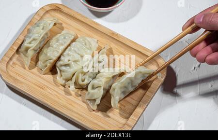 vista dall'alto della mano di un uomo con bastoncini di pollo su un tavolo di legno pronto a mangiare gyozas da un vassoio di legno, sano concetto di fast food orientale. Foto Stock