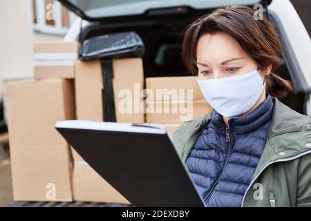 Servizio di consegna subappaltatore con maschera facciale controlla le consegne davanti di uno spazio di carico completo Foto Stock