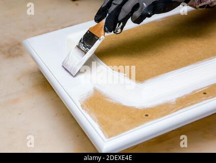 Riverniciatura porte armadio cucina con bianco gesso vernice in interni a casa. Dare alla vecchia cucina nuovo look concetto. Tenendo a mano uno strumento pennello. Foto Stock