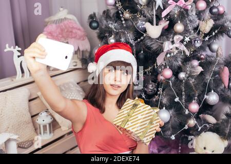 Adolescente ragazza in cappello di Santa prende selfie con regalo d'oro tra le decorazioni natalizie in rosa. Foto di alta qualità Foto Stock