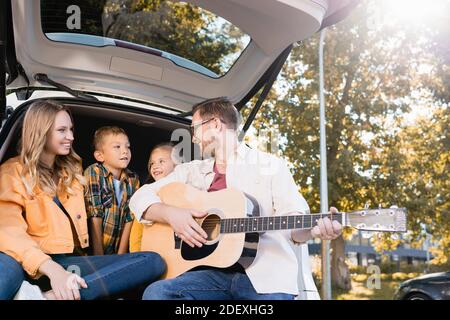 Sorridente padre che suona la chitarra acustica vicino a moglie e bambini dentro bagagliaio dell'auto Foto Stock