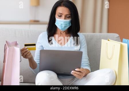 Signora matura in maschera medica utilizzando il computer portatile e la carta di credito Foto Stock