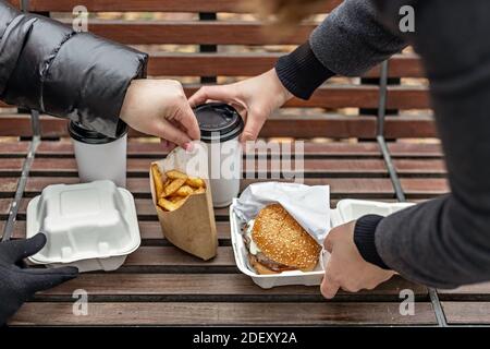 Le mani femminili stendono gli hamburger con tazze di caffè, le patatine fritte su una panca del parco. Fast food. Concetto di cibo da asporto. Foto Stock