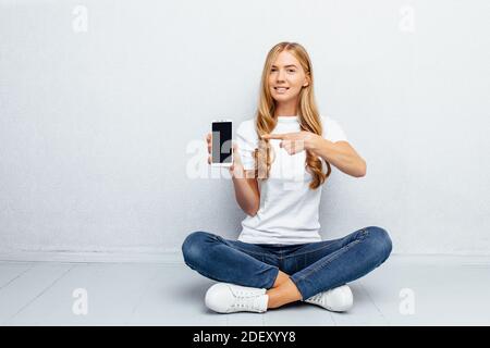 Una bella ragazza giovane che indossa una t-shirt bianca è seduta sul pavimento con le gambe incrociate che mostrano uno spazio vuoto schermo del telefono cellulare su sfondo grigio Foto Stock