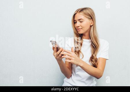 Giovane bella ragazza vestita di camicia bianca, tenendo il telefono e fissando lo schermo, seduto sul pavimento a gambe incrociate, isolato su sfondo grigio Foto Stock