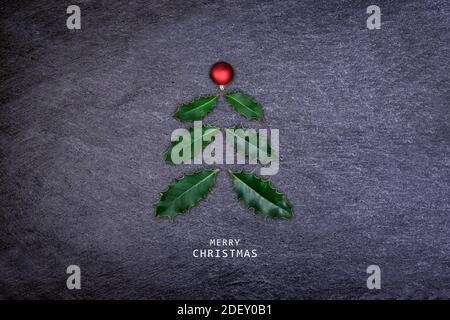 Albero di Natale fatto di foglie di agrifoglio e rosso bauble su pietra scura. Cartolina di Natale. Foto Stock