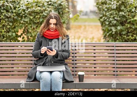 La giovane donna tiene uno smartphone e controlla i messaggi mentre è seduto su una panchina con un computer portatile in grembo Foto Stock