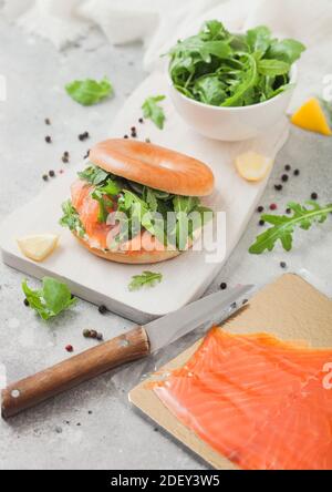 Panino fresco con bagel e salmone, formaggio cremoso e razzo selvatico su tavola bianca con pacchetto di salmone affumicato e coltello su sfondo chiaro. Foto Stock