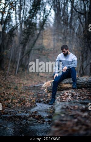 Giovane uomo seduto su un albero caduto appeso su un fiume nella stagione autunnale Foto Stock