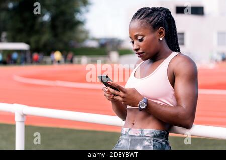 Foto d'inventario di un velocista afroamericano appoggiato su una rotaia della pista atletica utilizzando il suo telefono cellulare Foto Stock