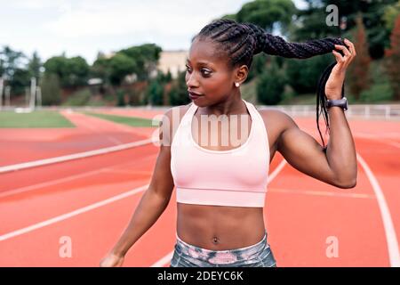 Foto d'inventario di una sprinter afroamericana in piedi su una pista atletica che tocca i suoi capelli intrecciati con la mano. Sport Foto Stock