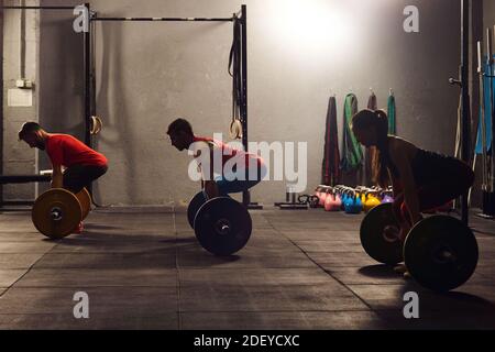 Foto di scorta di tre adulti in una palestra che si abbassa e solleva barbells. Indossano abbigliamento sportivo. Ci sono manubri sullo sfondo. Foto Stock