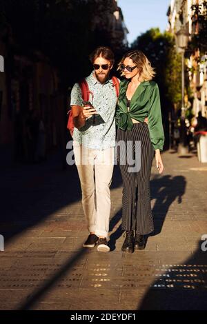 Foto in stock di una coppia nel loro 30 a piedi lungo una strada. Stanno guardando uno smartphone. Indossano un panno e occhiali da sole casual. Foto Stock