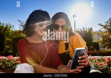 Foto di scorta di due ragazze adolescenti che guardano qualcosa su uno smartphone. Stanno sorridendo. Indossano un panno e occhiali da sole casual. Foto Stock