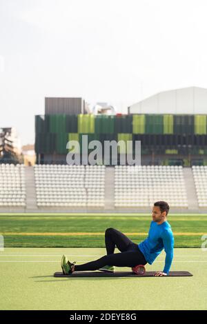 Foto d'inventario di atleta africano-americano seduto sulla pista che allunga le gambe con rullo in schiuma. Foto Stock