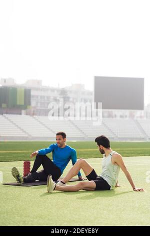 Foto di stock di atleti caucasici e neri di sesso maschile seduti in pista mentre si allungano le gambe e le braccia. Foto Stock